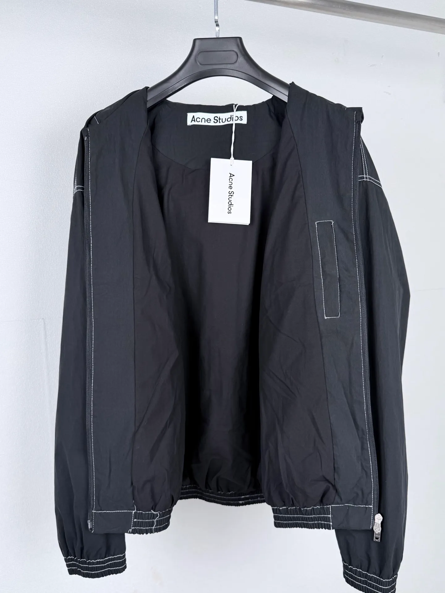 ACNE Studios back letter topstitch hooded jacket is made of fabric processing, contrasting topstitching, printing alignment and combination of elastic components. The fabric is treated with a physical wrinkle process, which does not rely on chemical coatings. It uses mechanical wrinkles to create a disordered texture effect on the surface of the fabric. While the fabric has a relaxed look, it still retains the structural tension of the woven fabric. During the cutting stage, the deformation caused by the folds of the fabric needs to be fully considered to ensure a stable silhouette after the pieces are spliced. The entire product uses contrasting topstitching as the main structural decoration. The seams of the cap, shoulder seams, cuffs, and hem are all exposed with contrasting topstitching. There are arc turns in different parts of the cut pieces. When sewing, it is necessary to control the fabric feeding rhythm and maintain a uniform stitch density. The elastic area of ​​the hem is sewn with multiple layers of materials. The layers of fabric, elastic bands, and hemming pieces are overlapped. It is easy for the line to shift and the fabric to arch, which requires high precision in the lathe operation. The Retro character printing on the back is the visual core of the entire garment. Multiple groups of characters are arranged in layers, giving it a worn and broken texture. The printing must ensure that the layer is strong and washable, but cannot cover up the details of the incomplete retro characters. At the same time, the printing positioning needs to be aligned with the middle seam of the back of the garment to avoid tilting and offset of the printing. The cuffs and hem are both elastic pleated structures. When sewing, you need to control the density of the fabric pleats to ensure a uniform and natural arc after shrinkage. The hood is spliced ​​into pieces and relies on multiple open threads to separate and shape it, ensuring that the hat is natural and three-dimensional and will not collapse against the face when worn. After all the sewing processes are completed, the garment needs to be ironed and finished to unify the wrinkles of the entire fabric and remove excess creases caused by the production process, finally presenting the complete Retro relaxed garment effect. Size: 46 48 50 52